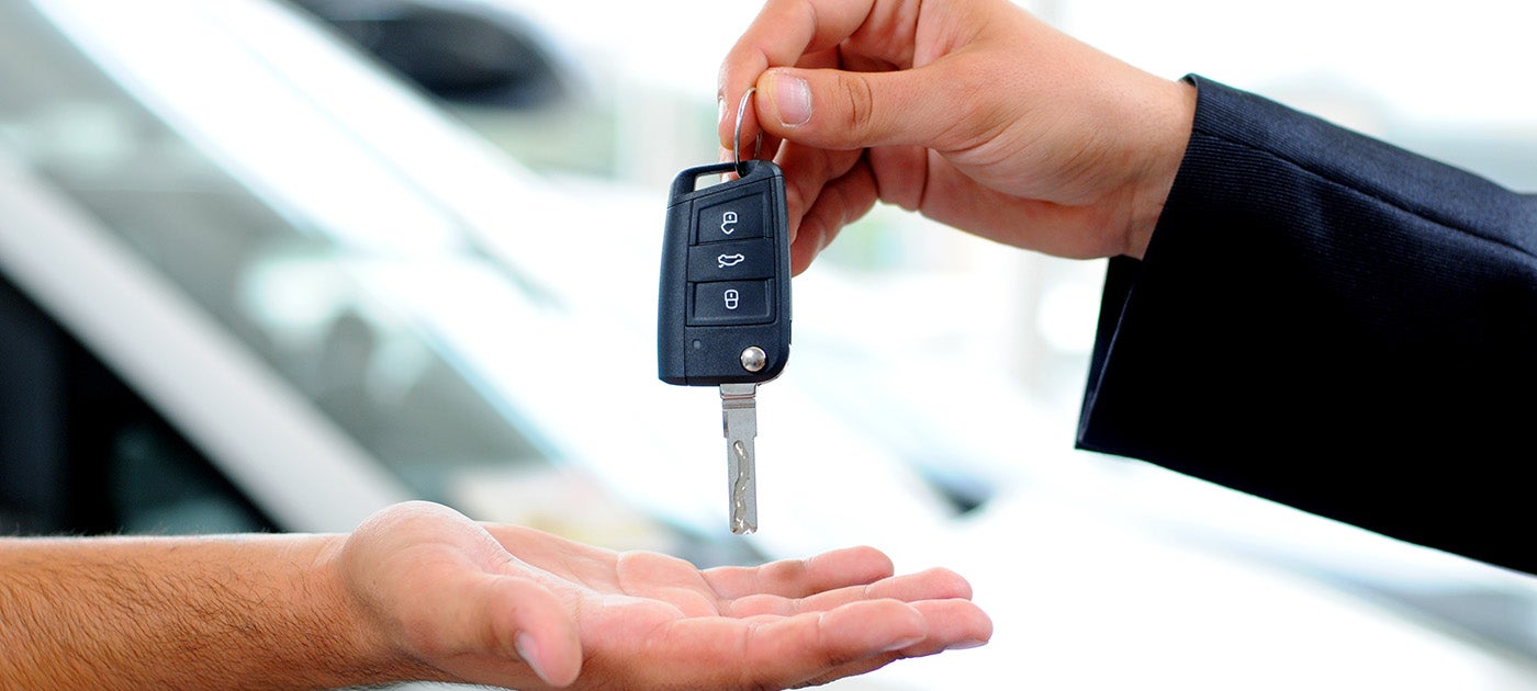 A hand in a dark suit jacket holding car keys over an outstretched hand, with a car visible in the blurred background.