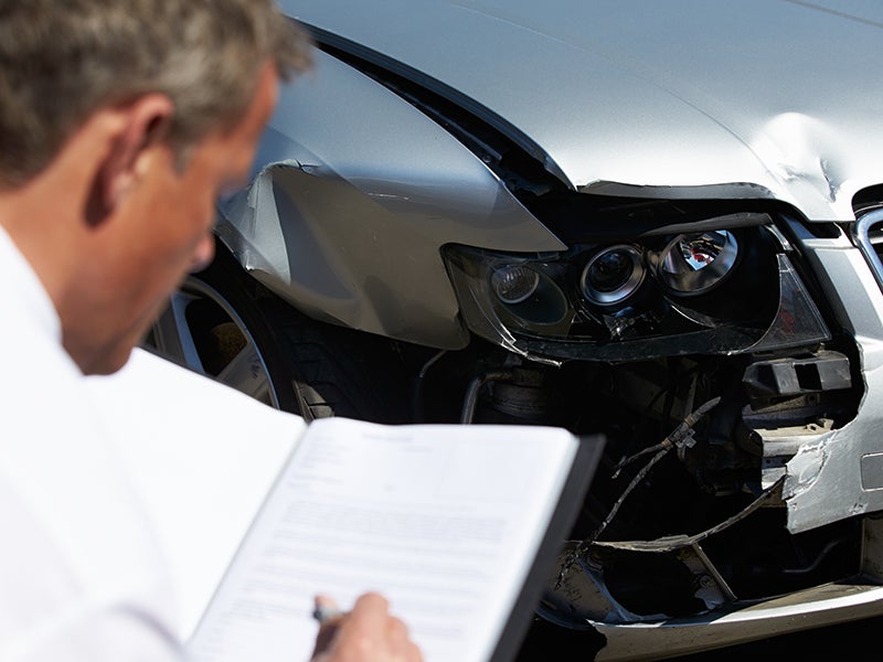 A person inspects a severely damaged grey car, likely after an accident, holding an open clipboard with papers.