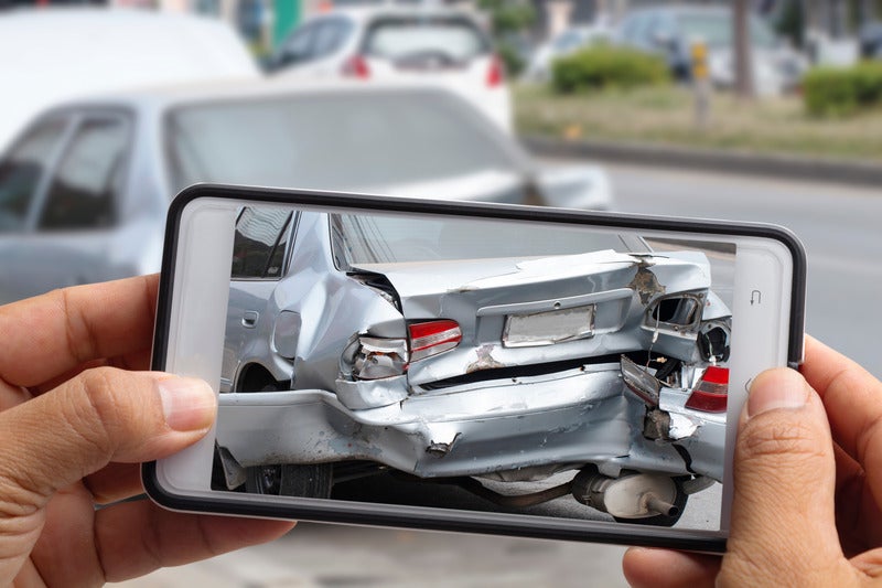 Hands holding a smartphone taking a photo of a silver car with severe rear-end damage.